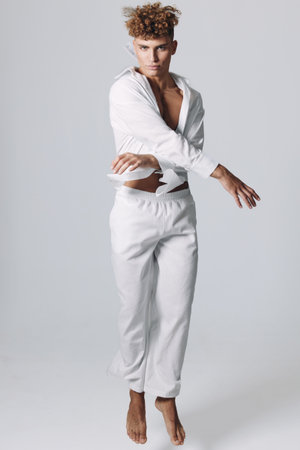 Young man with curly hair wearing white shirt and pants jumping in studio with neutral background,...