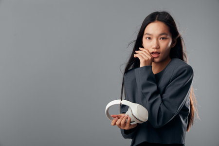 Young woman with long hair holding white headphones with thoughtful expression against gray background. Female portrait, casual wear and technology accessory for music listening or podcast.の写真素材