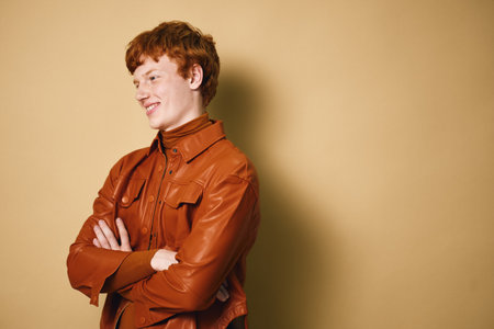 Young man in brown leather jacket posing with arms crossed, looking sideways smiling against beige...
