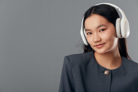 Young woman with long black hair wearing headphones and dark blazer smiling gently on gray background. Portrait, technology, lifestyle and music concept with casual elegance and youth culture.の写真素材