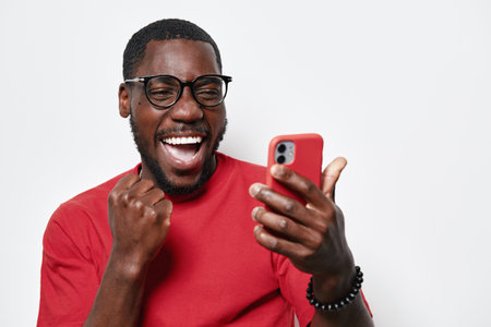 African American man with dark skin wearing glasses and red shirt celebrating with fist pump while looking at smartphone screen isolated on white background with joyful expression.の写真素材