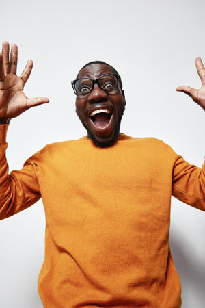 African man with dark skin and glasses wearing orange sweater expressing excitement with wide open mouth and raised hands on white background.の写真素材