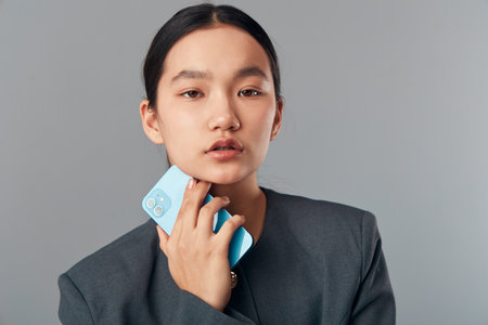 Young woman holding smartphone near face looking at camera with neutral expression wearing gray...