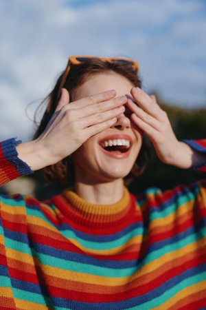 Woman smiling and laughing with hands covering eyes wearing colorful striped sweater and sunglasses...