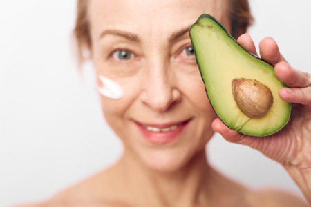 Smiling mature woman holds fresh avocado half near face with natural cream on cheek. Concept of...