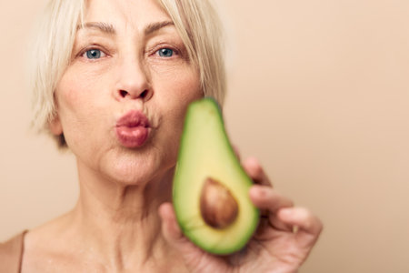Mature woman with short blonde hair holds half of avocado close to camera while puckering lips. Natural skin and healthy food concept with fresh fruit and positive expression.の写真素材
