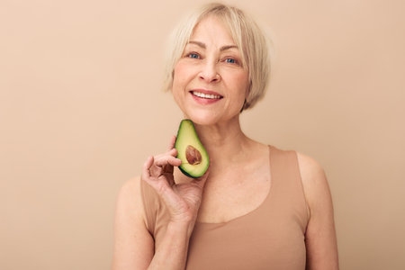 Smiling mature woman with short blonde hair holds half avocado near face on beige background....
