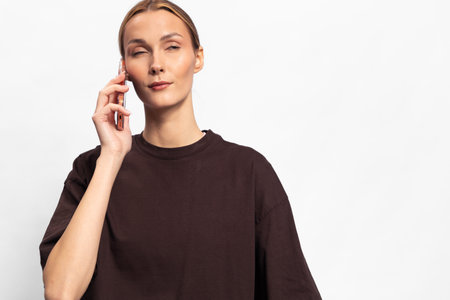 Young woman in dark loose shirt holds smartphone near ear with closed eyes. White background and...