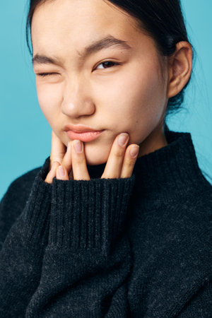 Young woman with dark hair wearing black sweater touches her chin in closeup portrait on blue background. Facial expression, casual and thoughtful mood with natural light.の写真素材