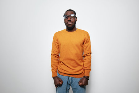 African American man with dark skin wearing orange sweater and glasses standing against white...