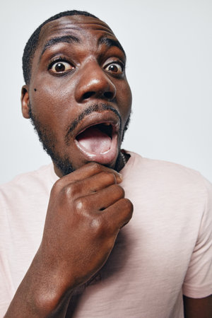 African American man with dark skin and short hair showing surprised expression with open mouth and hand on chin against white background. Casual clothing and emotional reaction.の写真素材
