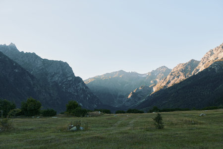 A tranquil alpine valley unfolds beneath rugged peaks as warm sunlight highlights a vast meadow,...