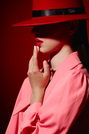 Fashion woman in a red hat and pink coat poses with softly lit face almost hidden in shadow against...