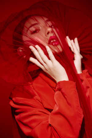 Fashion model in vibrant red coat and veil touches her face while posing dramatically. Evening makeup emphasizes glossy lips and emotive expression against monochrome background.の写真素材
