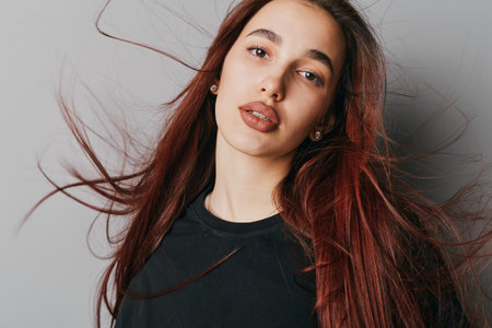 Young woman with long red hair and natural makeup posing in black shirt against gray background....