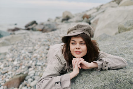 Young woman in casual clothes and hat lies on rocky beach, looking sideways with a relaxed smile....