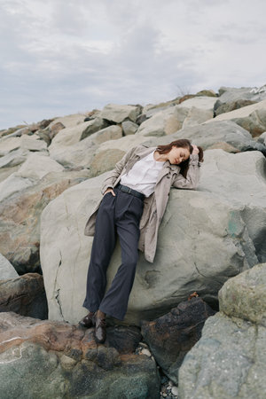 Young woman in coat and trousers leans against large rock on rocky beach under cloudy sky. Casual...