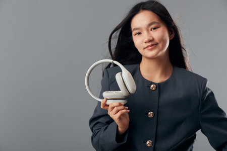 Young woman wearing dark jacket holds white headphones in hand smiling, portrait with long black...