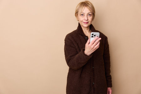 Middle aged woman in brown cardigan takes selfie with smartphone on beige background. Concept of...