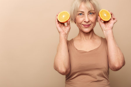 Smiling mature woman with short blonde hair holds two orange halves near face on beige background. Concept of healthy lifestyle, natural vitamins, skincare, and cheerful positive mood.の写真素材