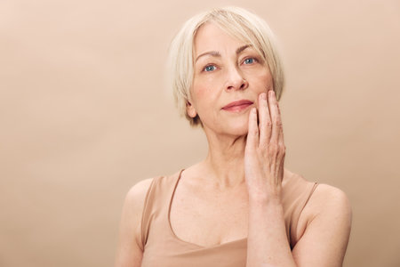 Mature woman with short blonde hair touches her face gently in a neutral beige background. Concept...