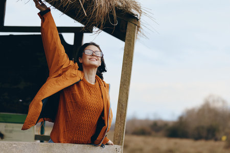 Happy woman in orange sweater and jacket enjoys outdoor lifestyle, smiling with arms raised near...