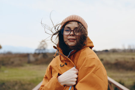 Young woman lifestyle outdoors wearing orange jacket and beige hat stands with wind blowing hair....