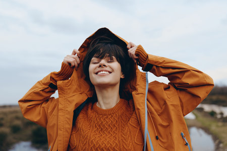 Happy woman in orange sweater and jacket enjoys outdoor lifestyle with eyes closed and smile....