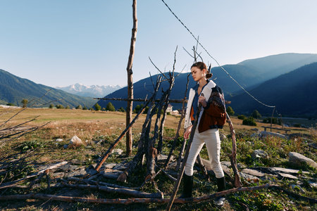 woman hiking with backpack walking through rural meadow near wooden fence with mountains in...