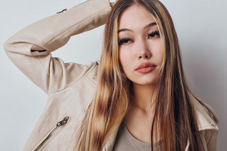 Young woman with long hair wearing beige jacket posing with hand behind head against white...