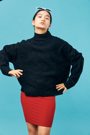 Young woman in black sweater and red skirt posing confidently with hands on hips against blue...