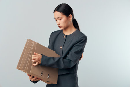 Young woman in dark formal jacket holding multiple cardboard folders with documents standing against...