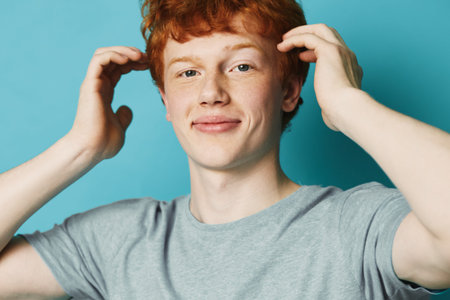 Young man with red hair and freckles wearing gray t shirt posing and smiling on blue background,...