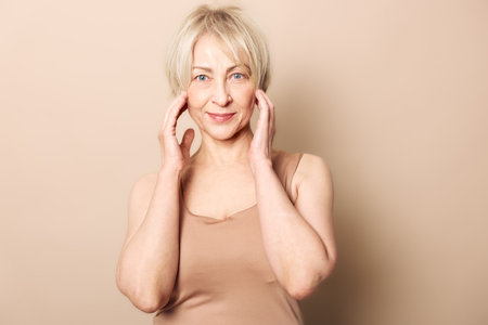 Mature woman with short blonde hair gently touching her face, smiling softly against beige...
