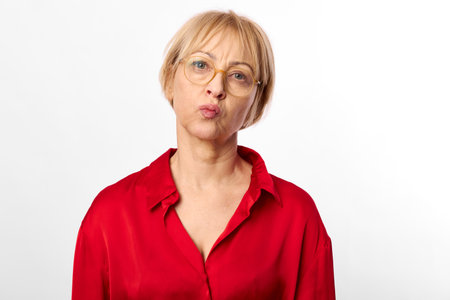 Mature woman with short blonde hair and glasses wears red shirt, looks skeptical with pursed lips on...