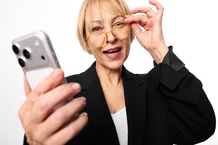 Middle aged woman in black jacket holding smartphone and adjusting glasses smiling at camera...