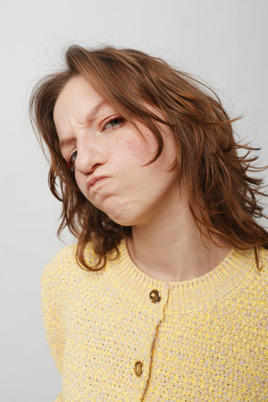 Young woman with messy brown hair wearing yellow sweater makes a displeased face with pursed lips...
