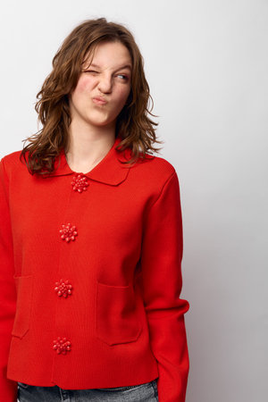 Young woman in red sweater makes funny face with squinted eyes and pursed lips against white...