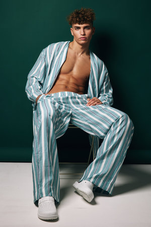 Young man wearing striped pajamas sitting in chair with hands in pockets and casual pose against green background. Relaxed shirt and shoes added leisure vibe to scene.の写真素材