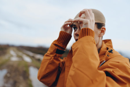 Young woman in orange jacket adjusts beige hat outdoors with blurred natural landscape in...