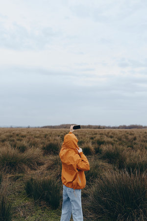 Woman lifestyle concept with a young woman in an orange jacket taking a photo on her smartphone in a natural field under a cloudy sky. Outdoor activity and casual fashion in nature.の写真素材