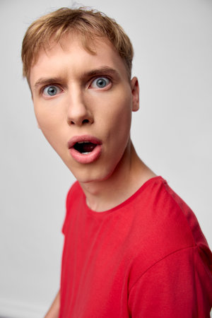 Surprised man in red shirt with wide eyes and open mouth, closeup expression captured in studio,...