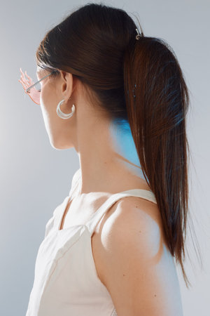Young woman with long dark hair in a white top and hoop earrings looks to the side. Studio portrait...