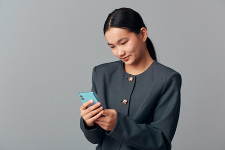 Young woman in dark jacket holding smartphone while smiling and looking at screen standing against...