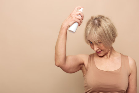 Mature woman with short blonde hair applies deodorant spray under her arm in beige tank top. Neutral...