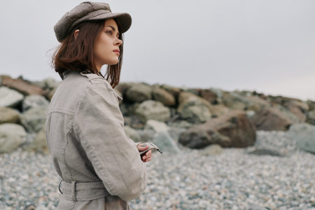 Young woman in beige coat and hat stands on rocky beach looking thoughtfully at distance under overcast sky. Concept of contemplation, autumn, and natural outdoor environment.の写真素材