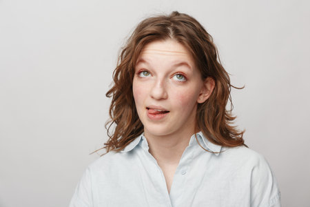 Young woman with curly hair and green eyes makes funny face sticking out tongue. Casual white shirt...