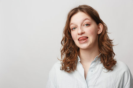 Young woman with curly hair wearing white shirt makes funny face sticking out tongue. Studio...