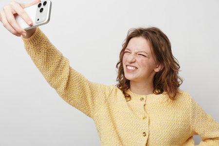Young woman in yellow sweater takes a selfie with smartphone, making a funny face. Studio shot on...