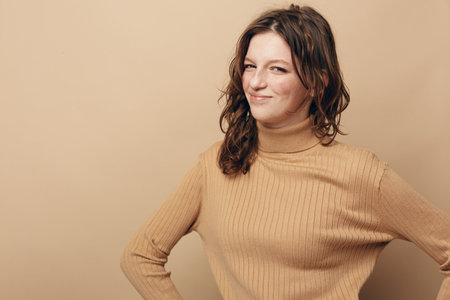 Smiling woman in beige turtleneck sweater poses confidently with hands on hips against plain...